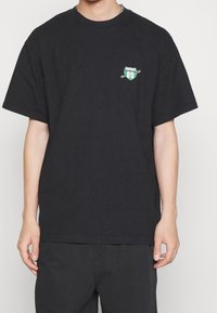 Camiseta de algodón negra con mangas cortas, que presenta un emblema gráfico en verde y blanco que dice "MP 89" en la zona del pecho izquierdo.