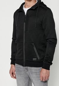 Felpa nera con zip, texture a coste, due tasche frontali e cappuccio con coulisse. Presenta una toppa con logo e dettagli della zip a contrasto.