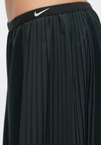 Czarna plisowana spódnica z elastycznym paskiem w talii i białym logo Nike. Materiał wydaje się lekki o gładkiej fakturze.