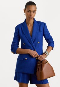 Kék, kétgombos blazer aranyló gombokkal és textúrával, hozzá illő sorttal és egy barna vödör táskával, amely arany díszítéssel rendelkezik.