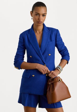 Modrá dvojradová blejzer s zlatými gombíkmi a textúrou, v kombinácii s ladnými šortkami a hnedou kabelkou typu bucket s zlatým detailom.