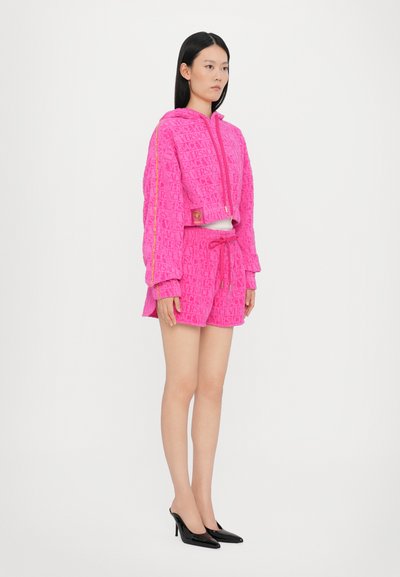 Roza krpana kapuca skrajšani komplet s šortami z vtisnjenim logotipom blagovne znamke. V kombinaciji s črnimi čevlji s konicami. Model stoji v profilu.