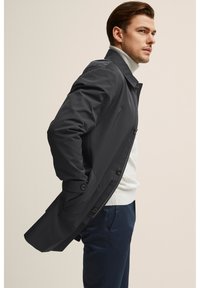 Musta vedenpitävä takki rakenteellisella suunnittelulla, jossa on korkea kaulus, napit ja sivutaskut, kerrostettuna kevyen neuleen päälle.