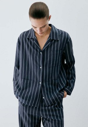 Person mit zurückgebundenem Haar, die ein dunkelblaues Nadelstreifenhemd und passende Hose trägt und mit den Händen in den Taschen nach unten schaut.