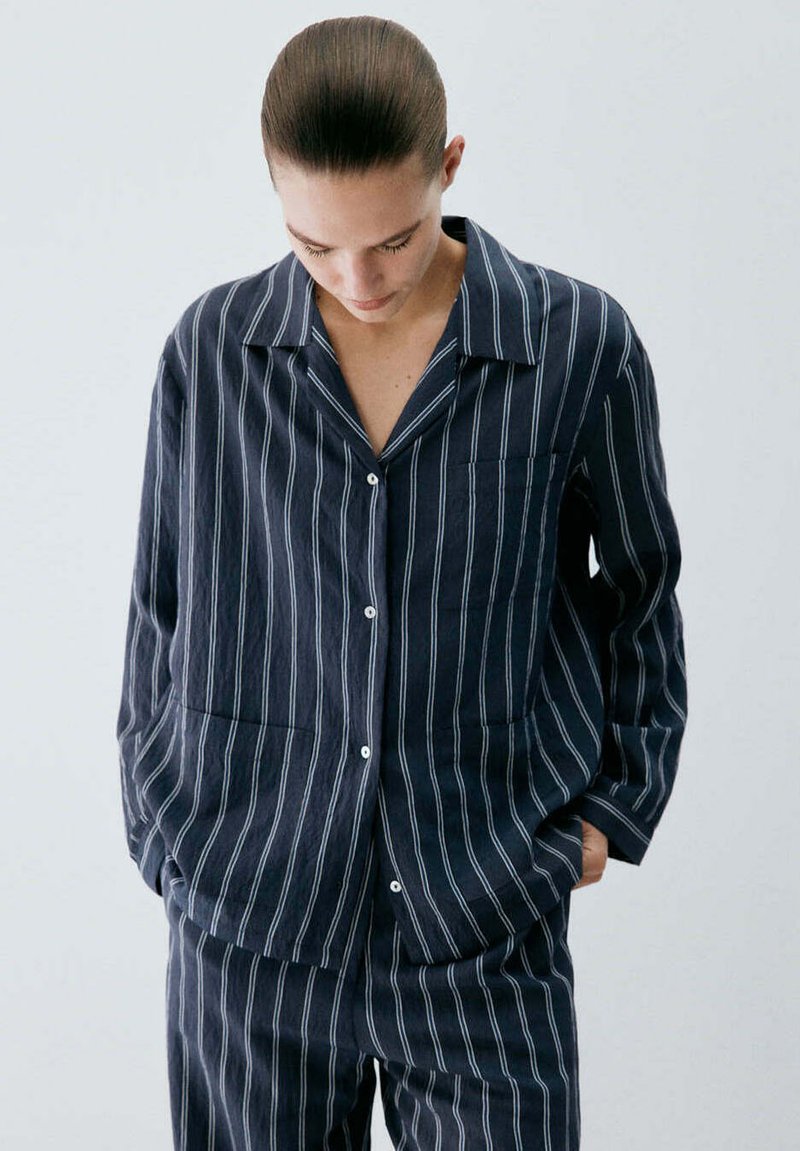 Personne aux cheveux attachés portant une chemise boutonnée à rayures bleu foncé et un pantalon assorti, regardant vers le bas avec les mains dans les poches.
