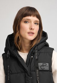 Czarna ocieplana kamizelka z kapturem, z naszywką z logo i akcentami z markowym paskiem, teksturowana tkanina oraz zapięcie na zamek.