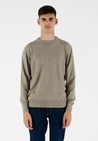 Beżowy sweter z okrągłym dekoltem, wykonany z miękkiej dzianiny, z długimi rękawami i ściągaczami. Noszony z ciemnoniebieskimi dżinsami.