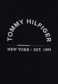 Tmavě námořnická látka s texturou. Na ní je tištěné bílé "TOMMY HILFIGER", které se klenutě vztahuje přes "NEW YORK • EST. 1985", s malou červenou a bílou akcentní linkou pod ním.