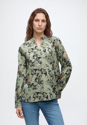 Femme aux cheveux bruns ondulés portant un chemisier à fleurs vert clair à manches longues et un jean bleu, debout devant un fond blanc uni.
