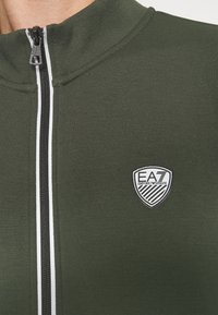 Mörkgrön zipjacka i slät tyg, med hög krage, svart dragkedja och ett vitt randigt accent längs framsidan. EA7-logotypen är synlig.