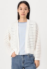 Joven mujer con el cabello negro lacio, vestida con un cárdigan blanco de punto abierto sobre una camiseta de rayas azules y blancas y jeans azules.