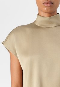 Top beige a collo alto realizzato in morbido tessuto di satin, con maniche corte a cappuccio e una vestibilità rilassata. Design minimalista senza motivi visibili.