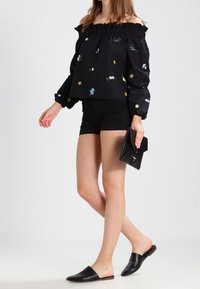 Zwarte top met open schouders en bloemrijke borduursels, gecombineerd met zwarte korte broek en platte instapschoenen. Houdt een zwarte clutch vast.