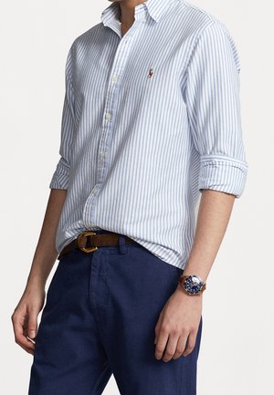 Mann trägt ein hochgekrempeltes blau-weiß gestreiftes Hemd, marineblaue Hose mit braunem Gürtel und eine Uhr mit blauem Zifferblatt und braunem Lederarmband.