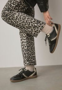 Schwarze und beigefarbene Sneaker mit Schnürdesign, ausgestattet mit strukturierten Paneelen, kontrastierenden Nähten und einer Gummisohle. Tragen mit gemusterten Leoparden-Hosen.