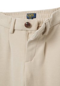 Pantalons pour enfants côtelés beiges avec une taille élastique, une fermeture à bouton et des détails de couture subtils pour une texture ajoutée. Étiquette de la marque OVS visible.