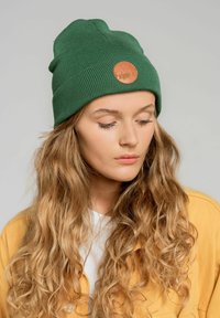 Zielona czapka ribbed z okrągłą brązową skórzaną łatką, noszona z żółtą kurtką i białą koszulą, na neutralnym tle.