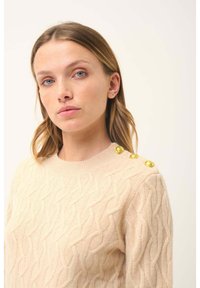 Maglione beige lavorato a maglia con motivo a trecce, colletto rotondo e tre bottoni dorati sulla spalla sinistra. Texture morbida e design aderente.