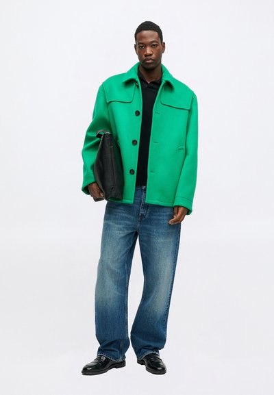 Chaqueta de mezcla de lana verde con cuello tipo solapa, botones negros, bolsillos inclinados, combinada con jeans de mezclilla azules y zapatos negros, sosteniendo un bolso negro.