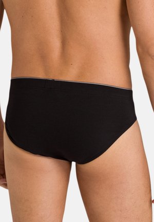 Mies, joka käyttää mustia lyhyitä alushousuja, näkyy takaa, korostaen istuvuutta ja vyötärönauhan yksityiskohtia.