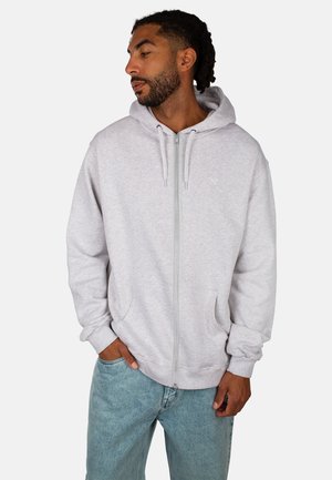 Mann mit geflochtenem Haar, der einen hellgrauen Reißverschluss-Hoodie und blaue Jeans trägt, mit einer Hand in der Tasche stehend und zur Seite blickend.