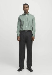 Cămașă butonată de culoare verde deschis, cu mâneci lungi, purtată cu pantaloni negri plisati și pantofi negri, stând pe un fundal alb.