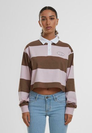 Chemise polo à manches longues courte rayée brun et rose avec un col blanc et un logo brodé. Un jean en denim est visible sous la chemise.