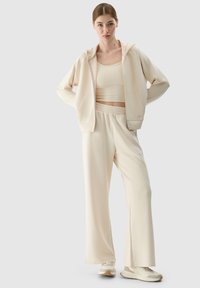 Conjunto de loungewear beige que incluye una sudadera con cremallera, una camiseta de tirantes ajustada y pantalones de pierna ancha, todo en una tela suave y lisa.