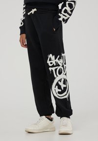 Czarne bawełniane dresy z elastycznym paskiem, bocznymi kieszeniami oraz białym nadrukiem w stylu graffiti, w tym uśmiechnięta buźka i tekst.