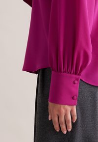 Blusa magenta con maniche lunghe a sbuffo, polsini a arricciati e due dettagli con bottoni, abbinata a un indumento grigio scuro sottostante.