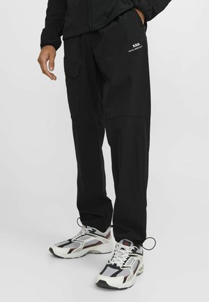 Black cargo pants with pockets, featuring a smooth texture and a relaxed fit. Branding in white on the thigh. Paired with black and white sneakers.