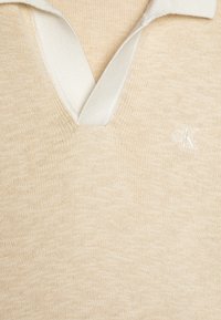 Polo in maglia beige con colletto a coste bianco, dotato di scollo a V e piccolo logo ricamato "CK" sul petto.