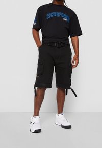 Osoba w czarnej koszulce z napisem "REDEFINED", czarnych szortach bojowych, czarnym pasku i białych sneakersach Nike stoi na jednolitym tle.