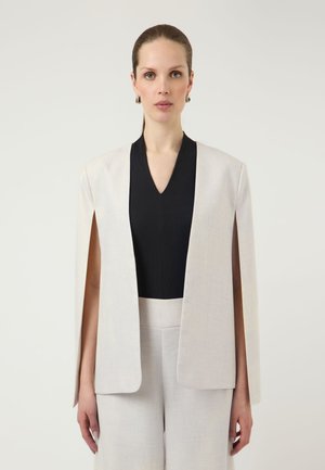 Woman wearing a black V-neck top under a light beige open-front cape blazer and matching wide-leg trousers, standing against a plain background.