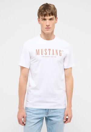 Junger Mann trägt ein weißes T-Shirt mit dem Aufdruck "MUSTANG Jeans Company Since 1932" und hellblaue Jeans, steht vor einem schlichten Hintergrund.