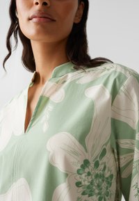 Light green floral-patterned blouse with a v-neckline and short sleeves, featuring white and darker green accents and smooth fabric texture.