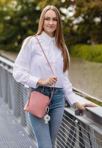 Růžová kožená kabelka přes rameno s hladkou texturou, kulatým tvarem a zipovým zapínáním, zdobená přívěskem připevněným k popruhu.