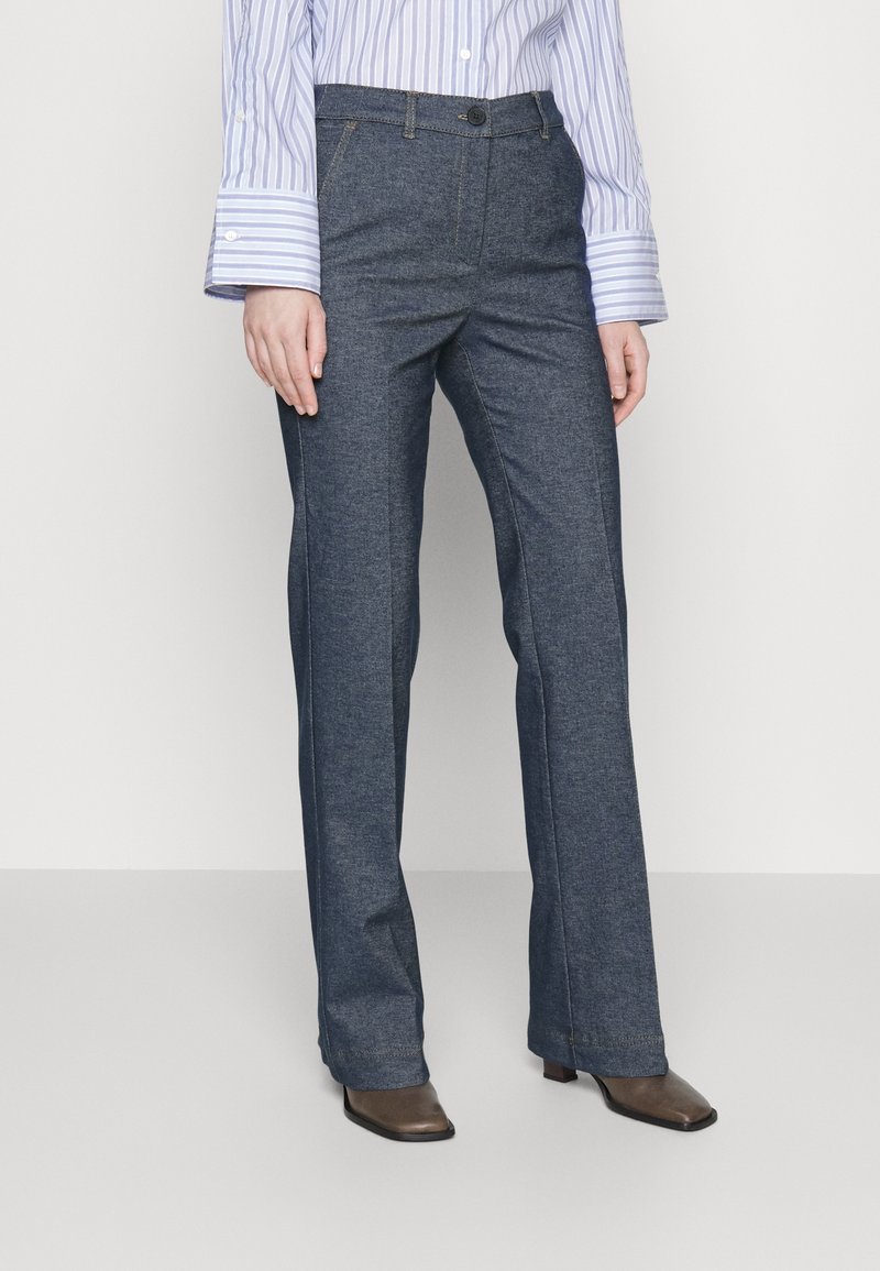 Žena v tmavě modrých rovně střižených kalhotách, hnědých botách se čtveratou špičkou a modro-bílé proužkované košili na knoflíky.