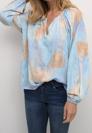 Kvinde iført en løs bluse med lange ærmer med blå og beige tie-dye mønster, kombineret med mørkeblå jeans.