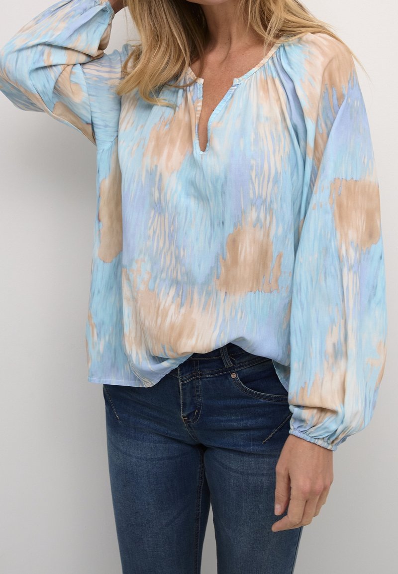 Femme portant un chemisier ample à manches longues avec un motif tie-dye bleu et beige, associé à un jean bleu foncé.