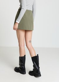 Bottes noires en cuir montant jusqu'aux genoux avec doubles boucles et semelles texturées, associées à une mini-jupe verte et un haut gris à manches longues.