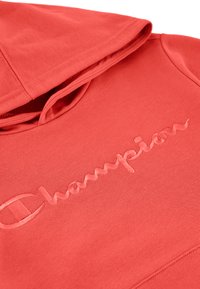 Felpa con cappuccio corallo realizzata in tessuto morbido. Presenta il logo "Champion" ricamato sul petto con dettagli testurizzati.
