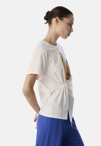T-shirt à manches courtes crème avec un détail noué à la taille, arborant une impression graphique colorée sur le devant. Associé à un pantalon plissé bleu.