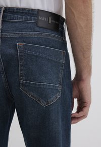 Dark blue denim jeans with a textured finish, featuring a rectangular leather label at the back, and visible stitching on pockets.