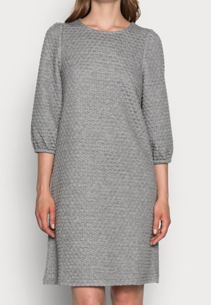 Kobieta ubrana w teksturowaną, szarą sukienkę za kolano z rękawami do połowy przedramienia i okrągłym dekoltem, stojąca na tle jednolitego białego tła.
