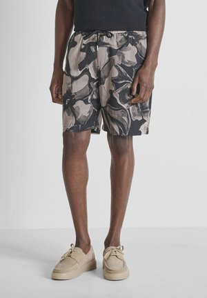 Uomo che indossa pantaloncini con stampa astratta neri e grigi, maglia nera e scarpe casual beige con lacci, in piedi su un pavimento uniforme.