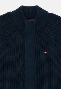 Cardigan en maille bleu marine avec un col haut côtelé, un tissu texturé et un panneau avant uni. Présente un petit logo en rouge, blanc et bleu.