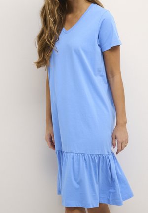 Femme portant une robe bleu clair à manches courtes et col en V avec un volant à l'ourlet, debout devant un fond clair uni.