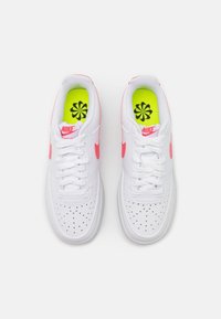 White athletic shoes with pink accents, perforated toe box, and white laces. Bright yellow insoles feature a black floral design.
