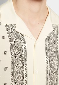 Chemise de couleur crème avec des motifs cachemire noirs et un col cranté au niveau du cou et du haut de la poitrine d’une personne.